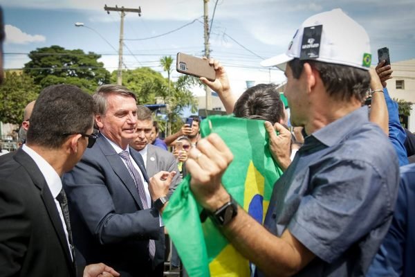 O presidente Bolsonaro durante motociata em Goiânia, onde também participou de evento evangélico. Ele olha para bandeira do Brasil que apoiador segura, cercado de multidão - Metrópoles