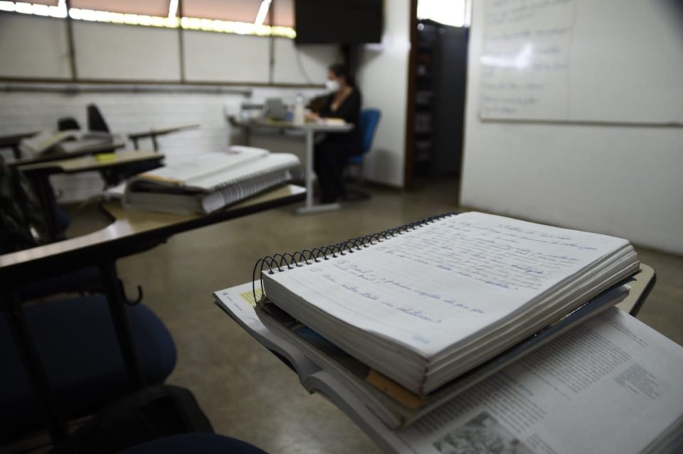 caderno em cima de carteira com professora na mesa em frente