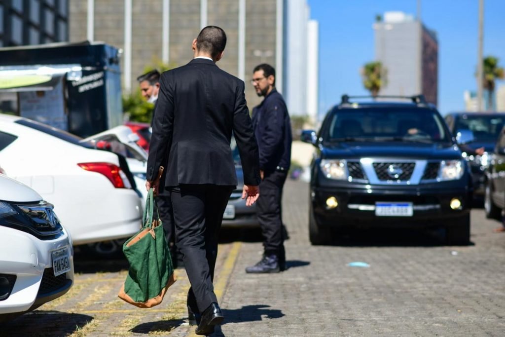 homens de preto no meio da rua com um carro preto na frente. um dos homens com uma bolsa verde na mão