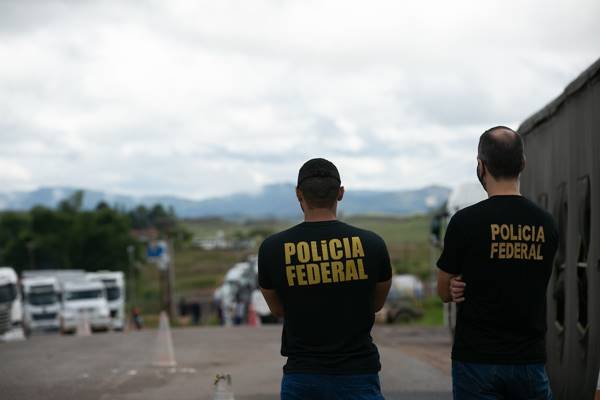 Fotografia colorida de dois policiais federais em pé de costas em estrada