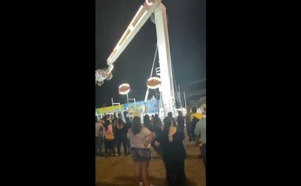 Pessoas observam brinquedo quebrado em parque de diversões de São Paulo onde pessoas estão presas de cabeça para baixo, de noite - Metrópoles