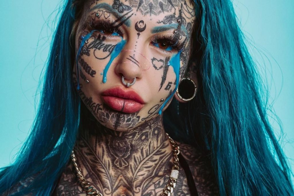 Mulher branca, com cabelos lisos e azuis, posando para foto em um fundo também azul. Ela tem quase todo o rosto tatuado e está com uma maquiagem que simula lágrimas, mas elas são densas e azuis.