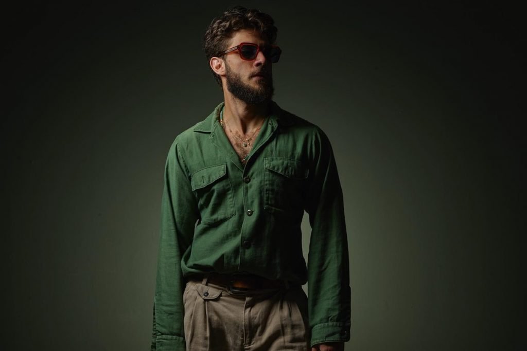 O ator Chay, um homem branco com cabelo e barba na cor castanha, posando para foto em um fundo escuro. Ele usa uma camisa verde, calça bege e óculos escuros.