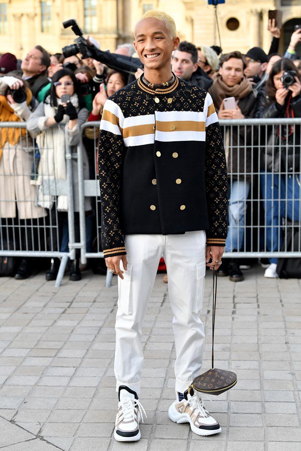 Jaden Smith na porta do desfile da Louis Vuitton, na Semana de Moda de Paris. Jaden é um jovem rapaz negro, com cabelo raspado descolorido, e usa uma camiseta preta, uma jaqueta preta com a logo da Louis Vuitton e botões dourados, e uma calça branca.