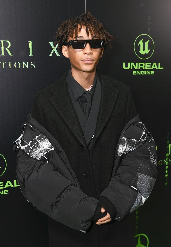 Jaden Smith, filho do Will Smith, no tapete vermelho da estreia do filme Matrix 4. Ele usa um look todo preto: camisa, blazer e um casaco estilo puffer, gigante, além de um óculos escuro pequeno.