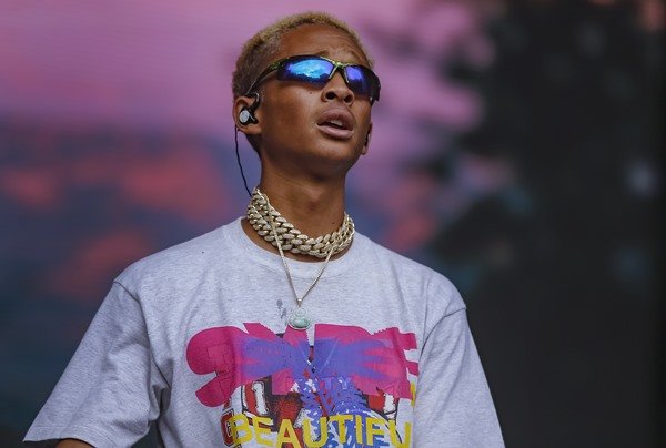 O ator e cantor Jaden Smith, filho de Will Smith, se apresentando no palco do Lollapalooza. Ele usa um óculos estilo juliete, que possui a lente espelhada, correntes douradas no pescoço e uma camiseta cinza com estampa gráfica rosa e amarela. 
