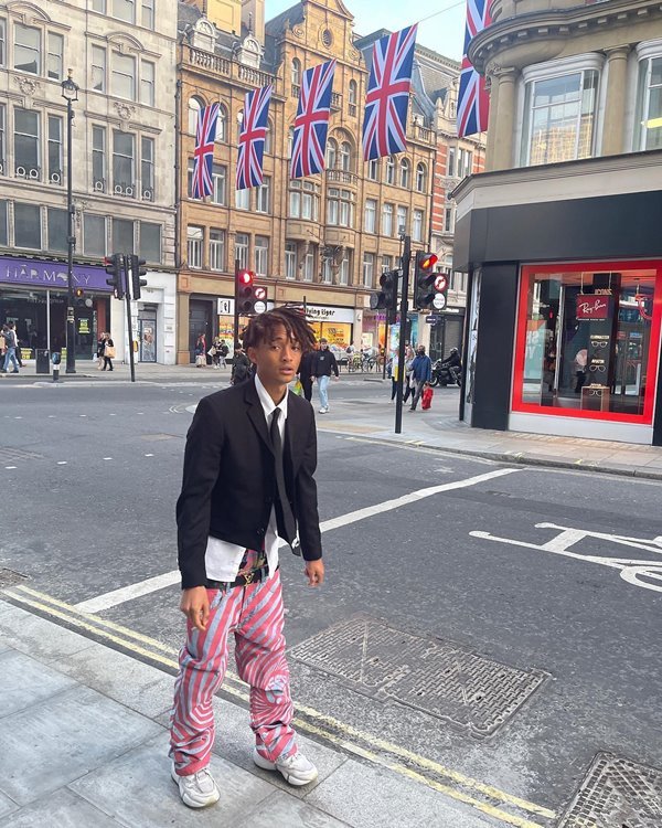 O ator e cantor Jaden Smith, filho de Will Smith, andando pelas ruas de Londres, na Inglaterra. Ele usa uma camiseta de botão branca, uma gravata e um blazer pretos, e uma calça estampada azul e rosa.