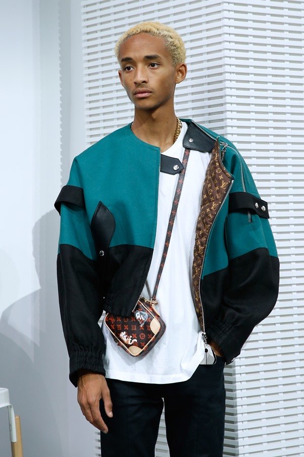 O ator e cantor Jaden Smith, um jovem rapaz negro com cabelos raspados e louros, posando para foto no Prêmio LVMH de 2018. Ele usa uma camiseta branca, um casaco moderno verde e preto e uma bolsa cruzada da Louis Vuitton.