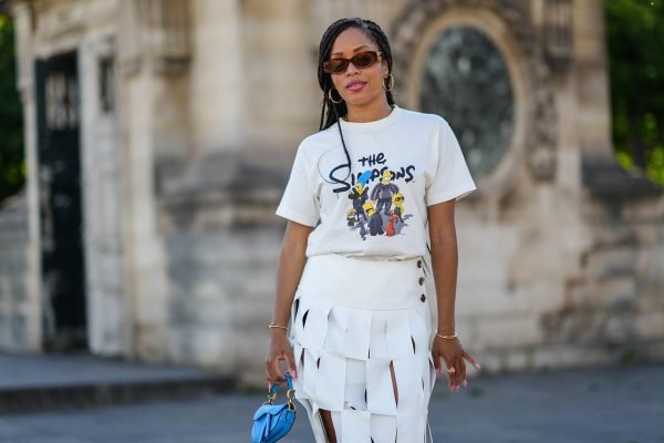 Mulher negra e jovem, com cabelos longos trançados, andando pelas ruas de Paris durante a semana de moda. Ela usa uma camiseta branca com uma estampa do desenho Os Simpsons, uma calça branca com recortes, uma mini bolsa azul e óculos escuros.