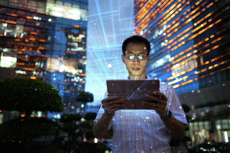 Foto colorida. Homem está com um tablet na mão em uma rua iluminada
