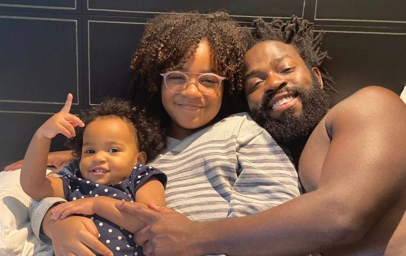 O ex-BBB22 e ator, Douglas Silva, posa para foto ao lado de sua filha pequena Maria Flor junto com sua outra filha bebê deitados em uma cama, sorrindo - Metrópoles