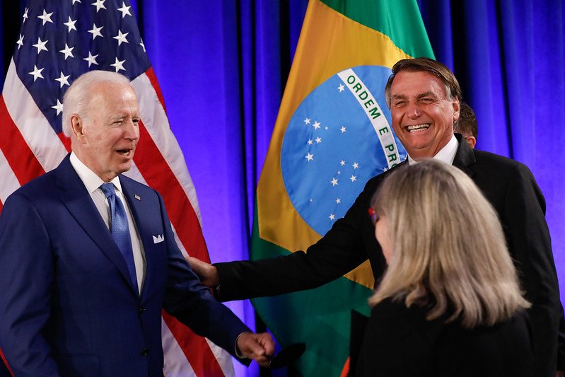 O presidente Bolsonaro cumprimenta o presidente americano Joe Biden na Cúpula das Américas, apertando as mãos enquanto uma mulher loira os olha, de costas.  Ao fundo, as bandeiras dos Estados Unidos e Brasil - Metrópoles