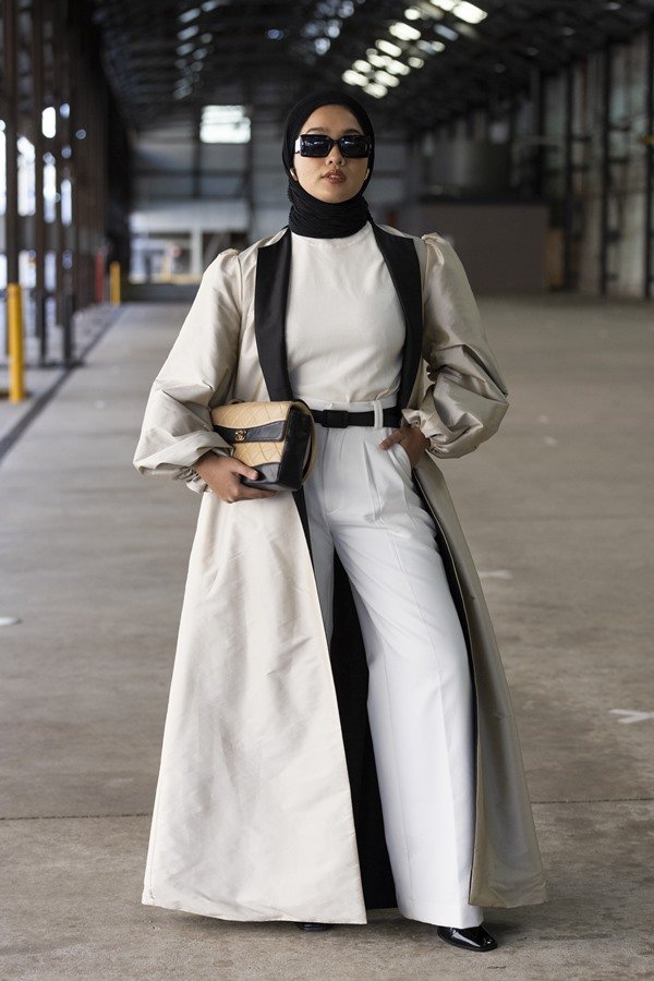Mulher morena posando para foto em um galpão vazio. Ela veste um turbante preto, tradicional da cultura islâmica; uma camiseta branca; calça branca com cinto preto; um casaco bege com detalhes pretos e uma bolsa nos mesmos tons, da marca Chanel.