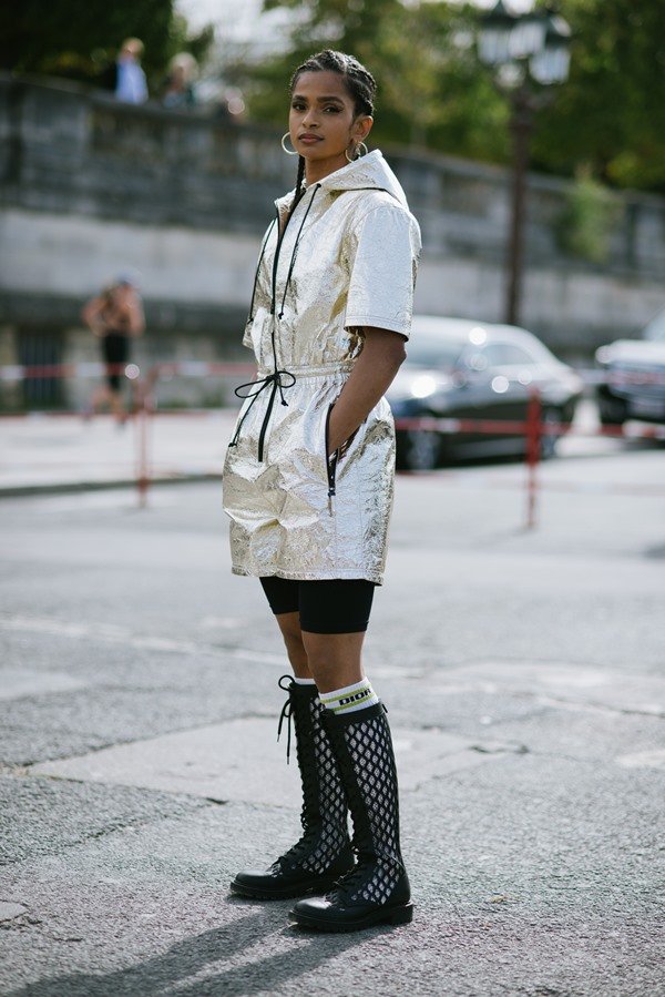 Mulher negra com cabelo trançado posando para foto na rua durante a Semana de Moda de Paris. Ela usa um macacão curto, dourado com detalhes pretos, meia branca longa da Dior e um coturno preto longo.