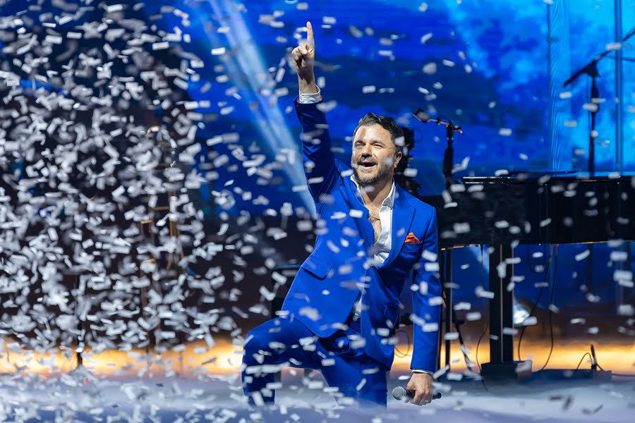 Cantor Mauricio Manieri em show no Vibra SP.  Ele está ajoelhado no, sorrindo e apontando pra cima em palco ao meio de confete - Metrópoles