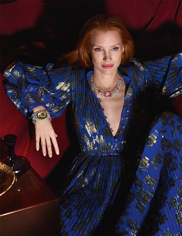 A atriz Jessica Chastain, uma mulher branca com cabelosos ruivos lisos e longos, posando para campanha publicitária de joias da marca Gucci. Na foto, ela usa um vestido azul com mangas bufantes, e detalhes brilhosos, e um colar e um brinco de diamantes com outras pedras preciosas.
