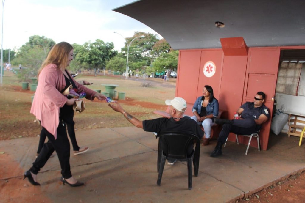 Mulher em pé entregando papel para homem sentado de costas. Duas outras pessoas estão sentadas, encostadas em posto médico no parque