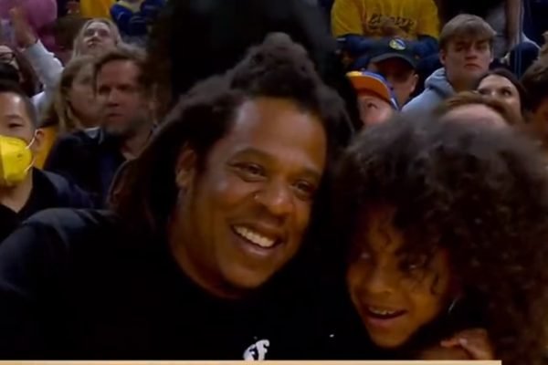 O rapper Jay-Z abraça sua filha com a cantora Beyonce, Blue Ivy, durante jogo de basquete de NBA. Ambos sorriem e é possível ver várias pessoas atrás nas arquibancadas - Metrópoles