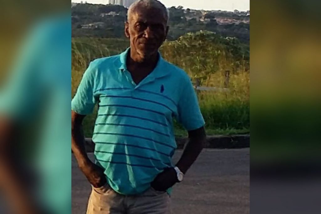 Homem com cabelos brancos, pele escura usando blusa azul e bermuda bege