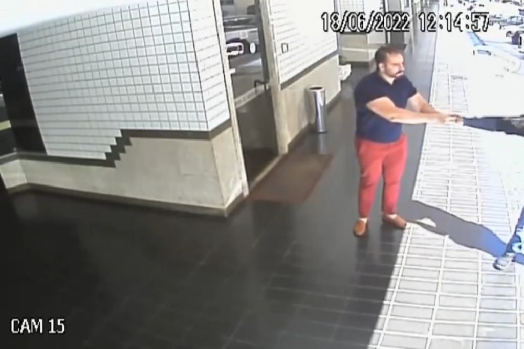 homem de camisa azul e calça caqui entrega relógio para assaltante em pilotis do prédio