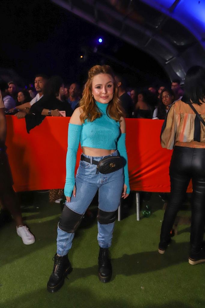 Na foto, mulher de blusa de manga comprida azul, com recorte nos ombros e calça jeans com detalhes pretos no joelho posa para a câmera. Ela também usa botas
