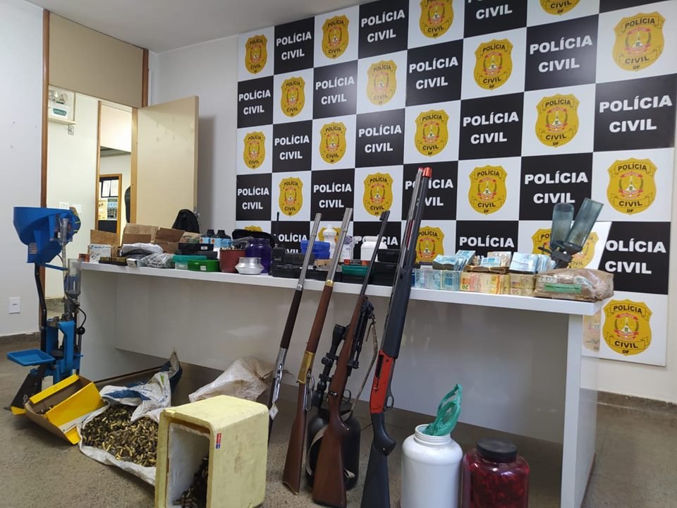 Armas encostadas em mesa branca com objetos e painel da Polícia Civil do DF ao fundo