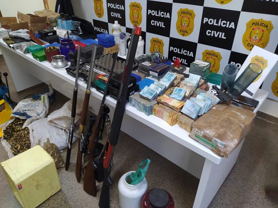 Armas encostadas em mesa branca com objetos e painel da Polícia Civil do DF ao fundo