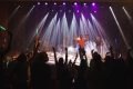 homem de blusa laranja canta em palco com banda e publico aparece em silhueta na frente