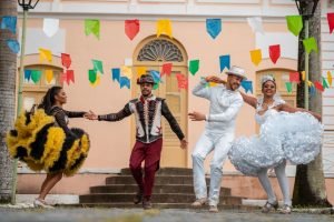 Casais vestido de noivos com roupas típicas juninas dançam sorrindo frente à casarão cor de rosa e rua enfeitada com bandeirinhas - Metrópoles