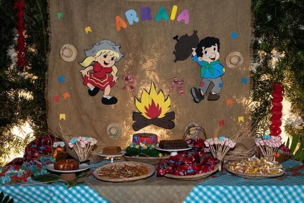 Mesa com diversos práticos típicos de São João frente a banner com desenho de casal dançando próximo de fogueira escrito "Arraia" - Metrópoles
