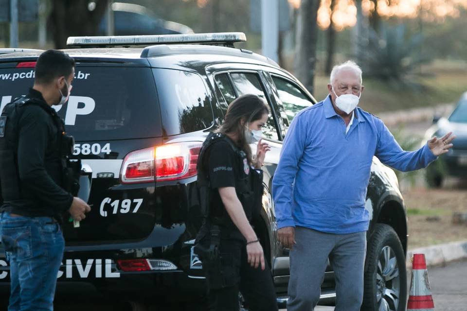 foto colorida de homem de blusa azul e máscara facial cercado por policiais