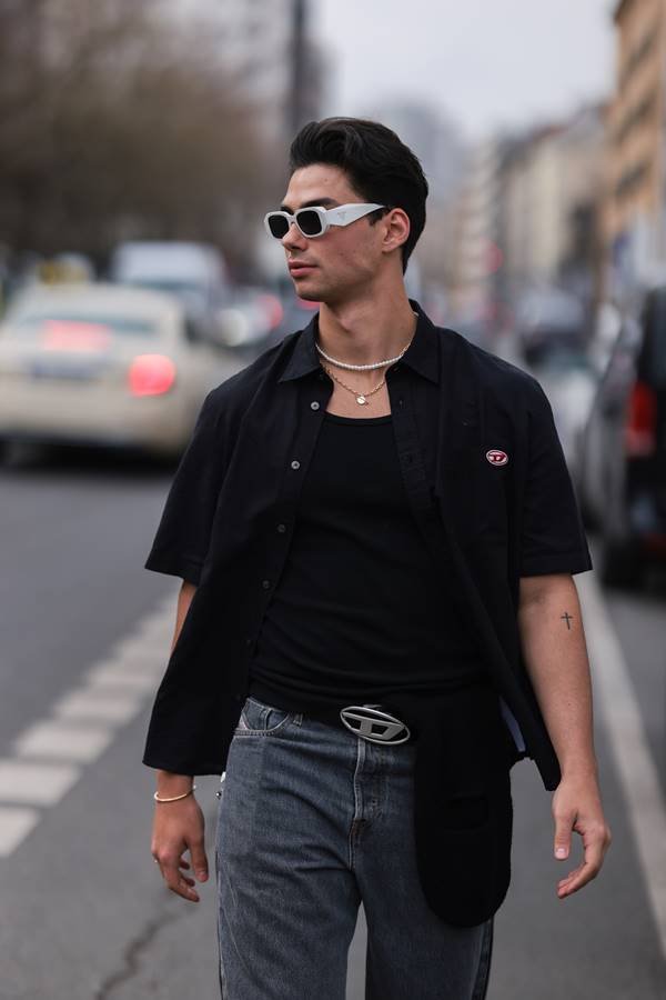 No street style, homem branco de cabelo escuro. Ele usa camiseta preta com sobreposições de camisa, além de óculos escuros e colar de pérolas discretas