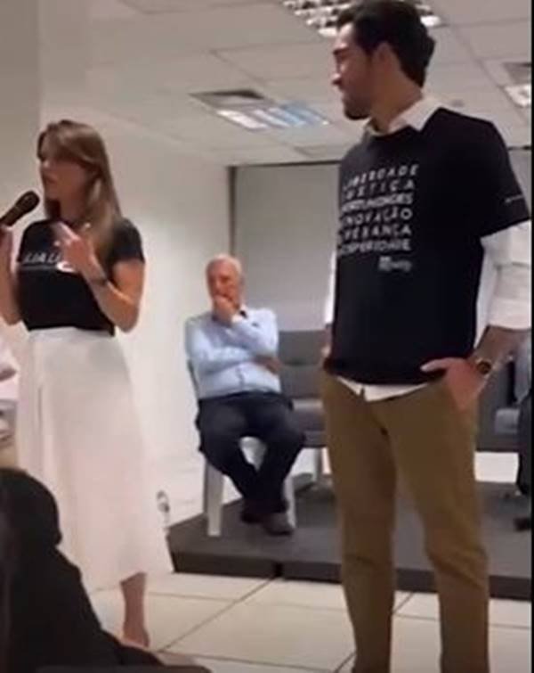 Fotografia colorida de mulher de blusa preta e saia branca segura microfone ao lado de homem de camiseta preta
