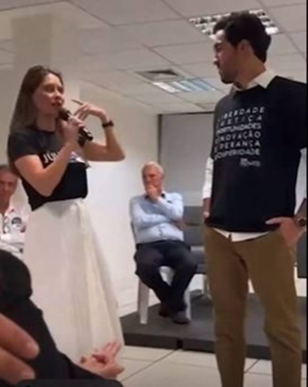 Fotografia colorida de mulher de blusa preta e saia branca segura microfone ao lado de homem de camiseta preta