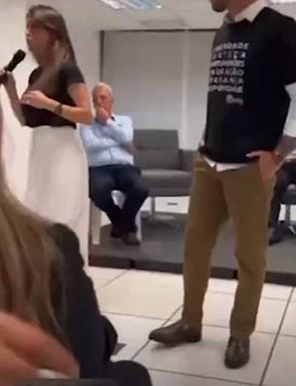 Fotografia colorida de mulher de blusa preta e saia branca segura microfone ao lado de homem de camiseta preta