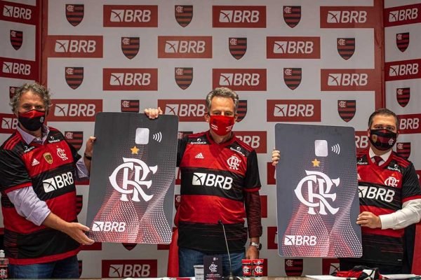 Três homens com uniforme do Flamengo vermelho e preto em pé segurando cartões grandes com símbolo do time. Atrás deles há um banner do Flamengo