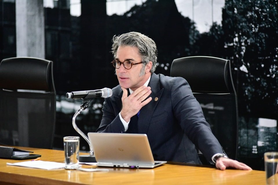 Homem branco de terno, com cabelos grisalhos e óculos sentado em cadeira preta falando em microfone. Ele coloca uma mão próximo a garganta e a outra na mesa. Há um notebook e um copo com água na mesa