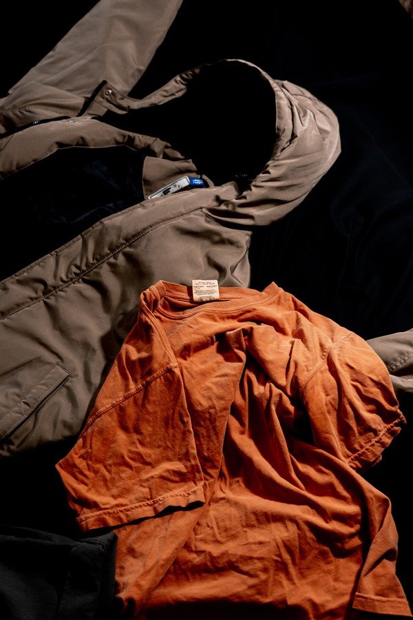 Casaco acolchoado marron, com capuz, e camiseta caramelo, ambas as peças jogadas no chão escuro.