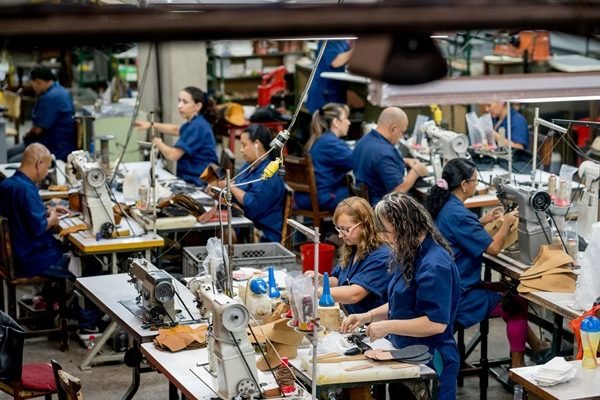 Mulheres e homens trabalhando em uma fábrica de roupas. Eles mexem em máquinas de costura e vestem, todos, um uniforme que é um macacão azul reoneração