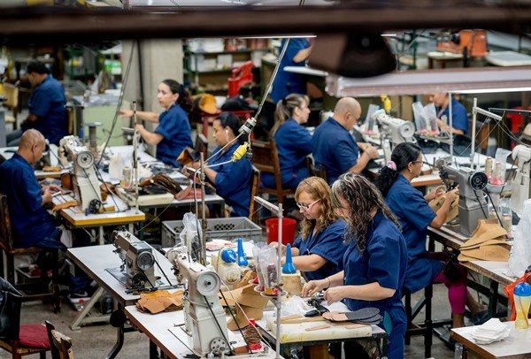 Mulheres e homens trabalhando em uma fábrica de roupas. Eles mexem em máquinas de costura e vestem, todos, um uniforme que é um macacão azul