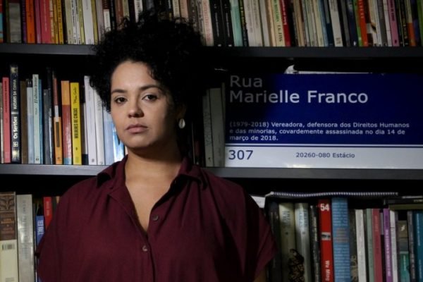 A jornalista Bianca Santana posa em frente a uma estante com uma placa de rua que traz o nome da vereadora Marielle Franco