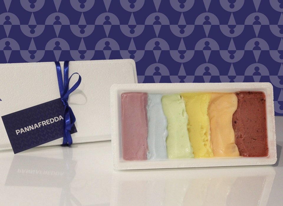 Sorvetes coloridos em pote branco. Tampa com fita azul escuro do lado. O fundo da imagem é azul com desenhos