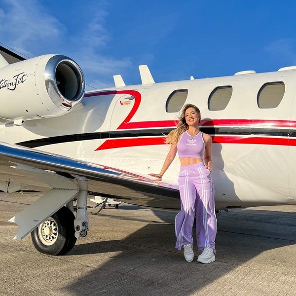 A influenciadora e esposa do cantor Zé Felipe, Virginia, posando para foto encostada em um avião particular na cor branca com listras vermelhsa e azuis. Ela veste um conjunto da Adidas de top e calça folgada na cor lilás com listras brancas.