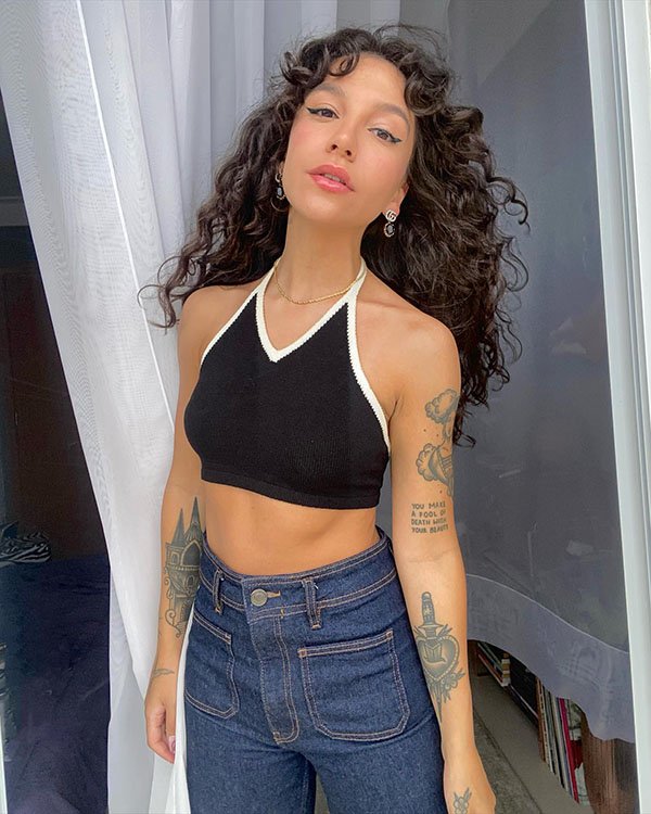A cantora Priscilla Alcantara, uma mulher jovem de pele branca, tatuagens, e cabelo cacheado longo, posando para foto. Ela usa um top curto preto com costura branca e uma calça jeans de cintura alta.