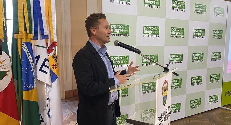 Homem de cabelos escuros, pele branca falando em frente a um microfone