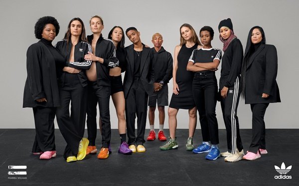 O cantor Pharrell Williams, um jovem negro, magro e com cabelo curto e descolorido, posando para foto em campanha da Adidas. Ao seu redor, várias mulheres de diferentes idades, raças e corpos usam roupas da marca esportiva