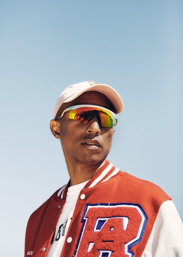 O cantor Pharrell Williams, um jovem negro, magro e com cabelo curto, posando para foto ao ar livre. Ele veste uma camiseta branca, uma jaqueta vermelha, óculos furta-cor no modelo juliet e um boné rosa. Todas as peças são da Adidas