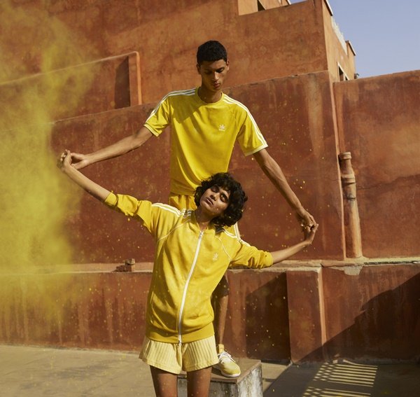 Campanha de divulgação de uma coleção do cantor Pharrell Williams com a marca Adidas. Na foto, uma mulher e homem, ambos jovens, de pele clara e cabelo preto, parecem estar dançando em roupas amarelas com listras brancas da marca