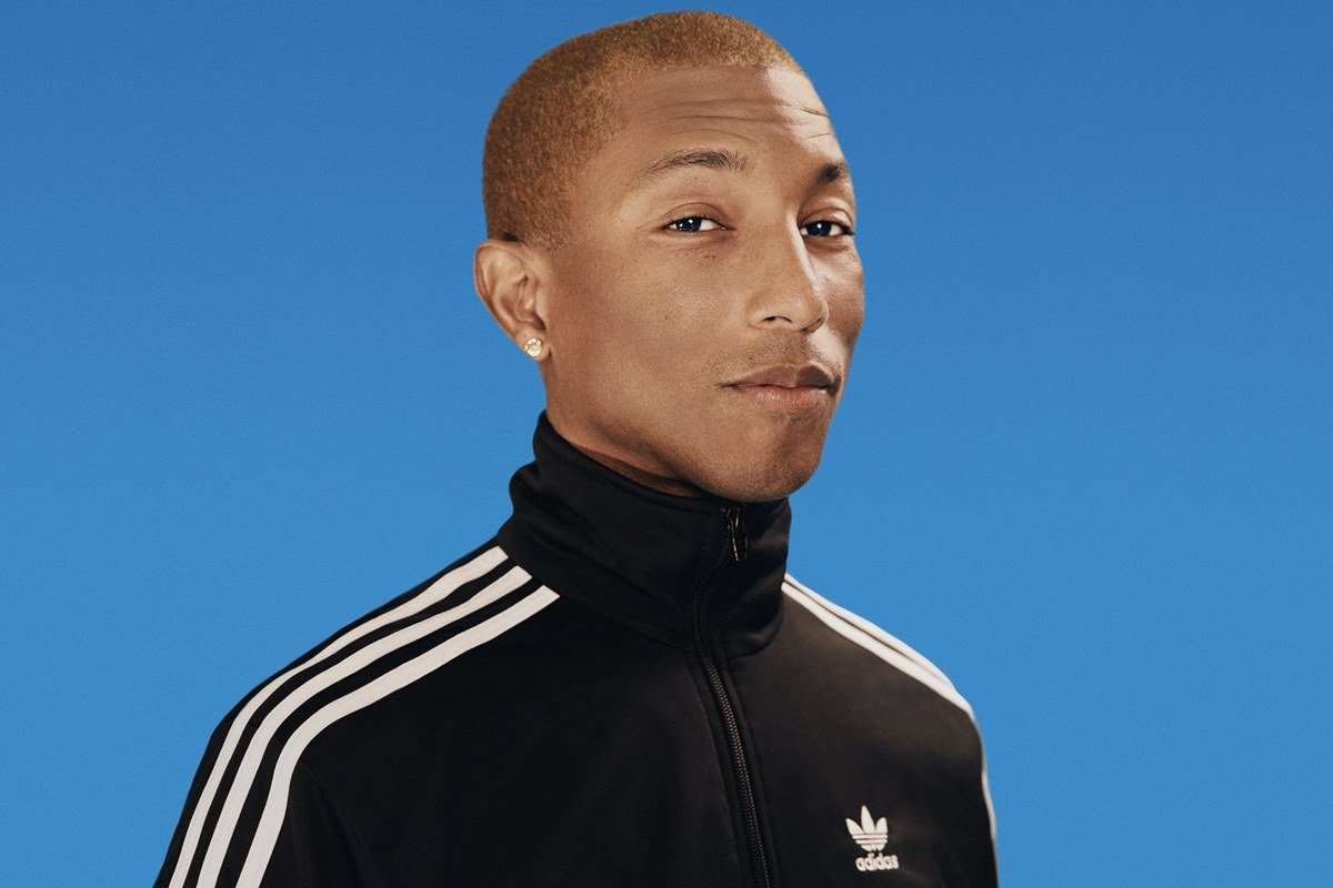 O cantor Pharrell Williams, um jovem negro, magro e com cabelo curto, posando para foto em um estúdio que possui fundo azul. Ele veste um casaco de moletom preto com listras brancas da Adidas
