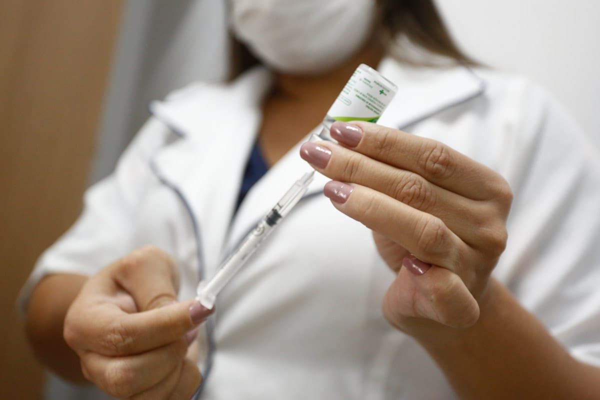 imagem colorida mostra mulher usando jaleco branco e máscara segurando seringa e vidro de vacina - Metrópoles